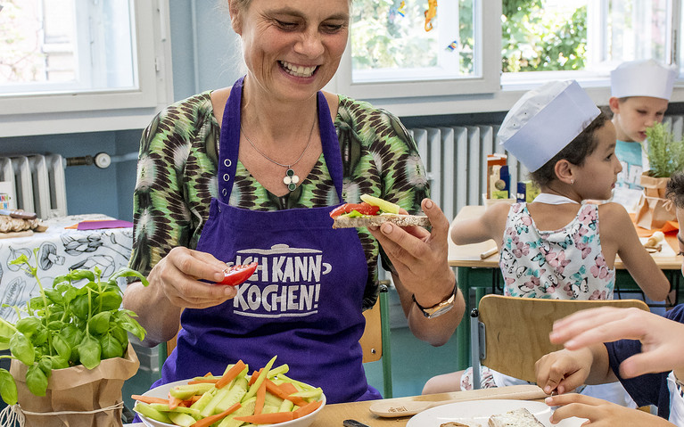 Stiftungsgründerin Sarah Wiener sitzt gemeinsam mit Kindern im Rahmen einer Ich kann kochen!-Kochaktion am Tisch und isst das selbst gekochte Essen.