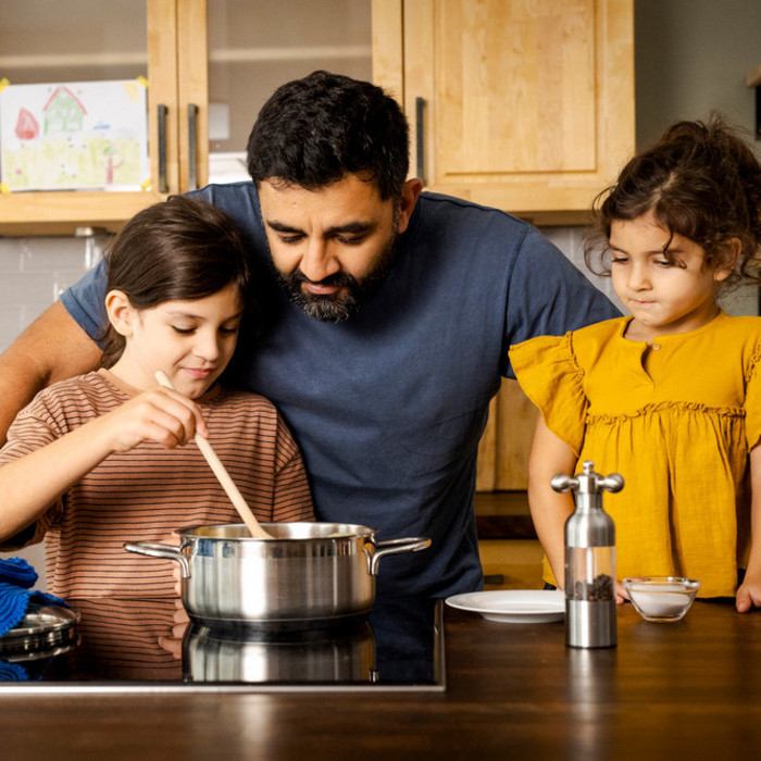 Vater und zwei Töchter stehen in der Küche und schauen in einen Kochtopf